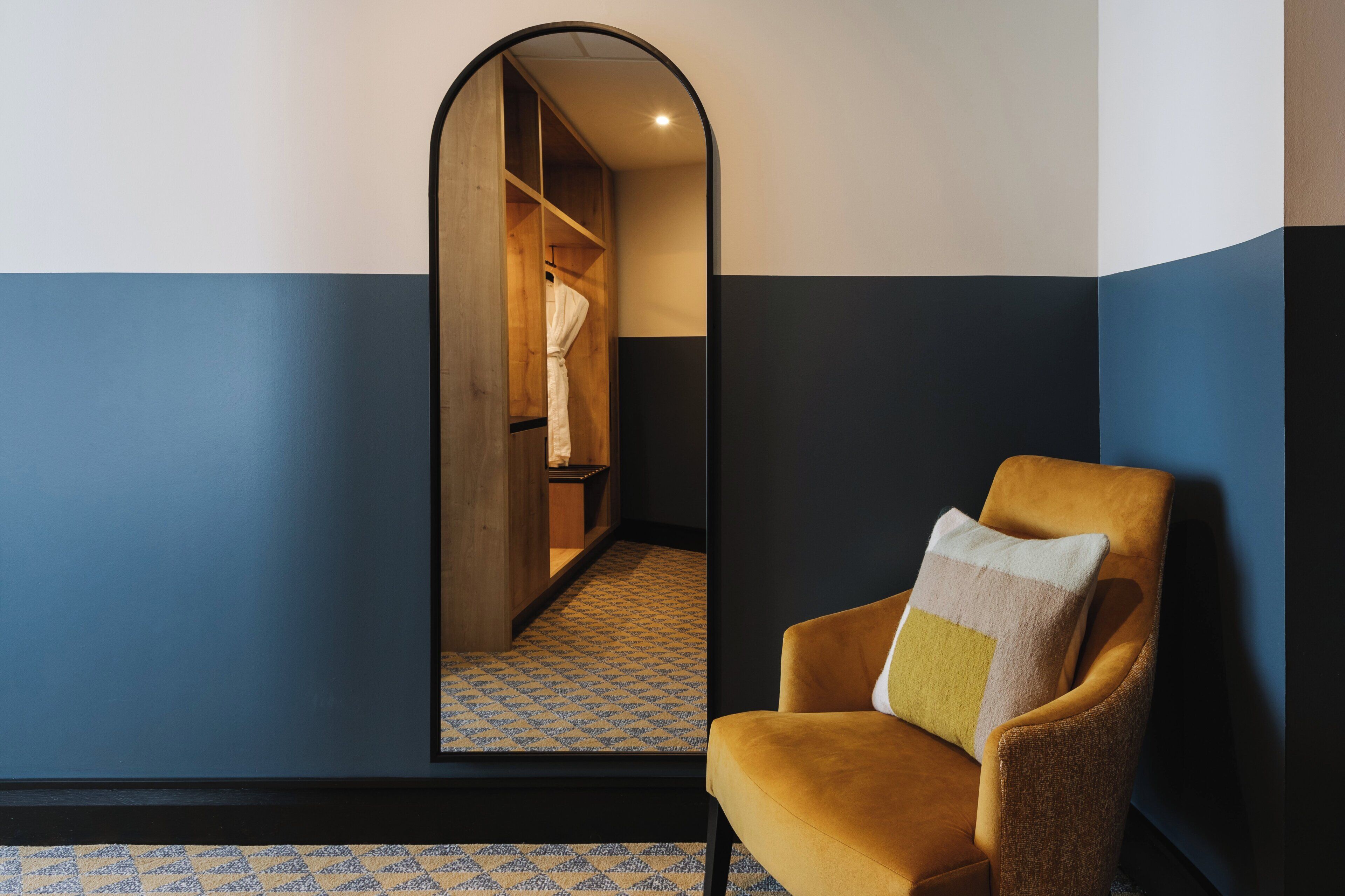 A room with a mirrored wall, a wooden wardrobe, a patterned floor, and a yellow armchair with a cushion.