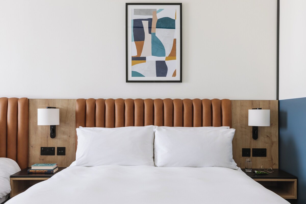 Een hotelkamer met een houten hoofdbord, witte beddengoed, twee nachtkastjes met lampen en een lijst met een abstract schilderij aan de muur.