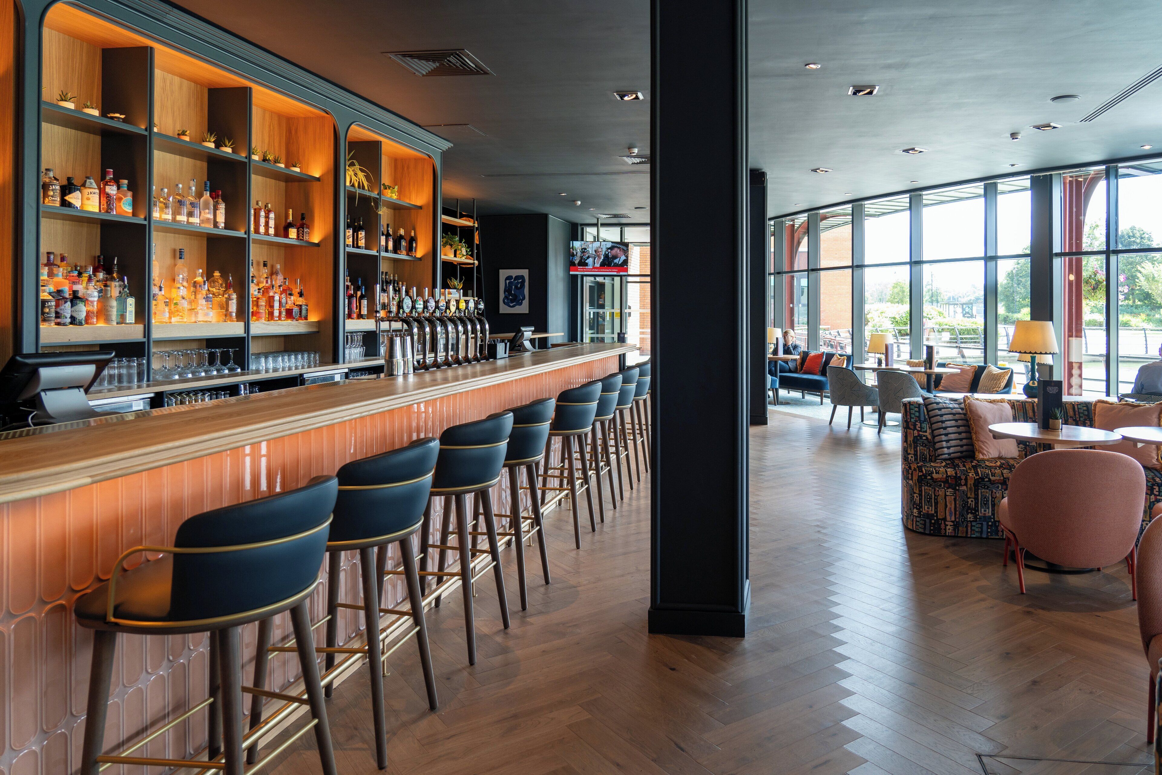 Una zona de bar con varios taburetes, una barra repleta de botellas y un área de estar con mesas y sillas.