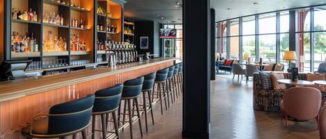 Una zona de bar con varios taburetes, una barra repleta de botellas y un área de estar con mesas y sillas.