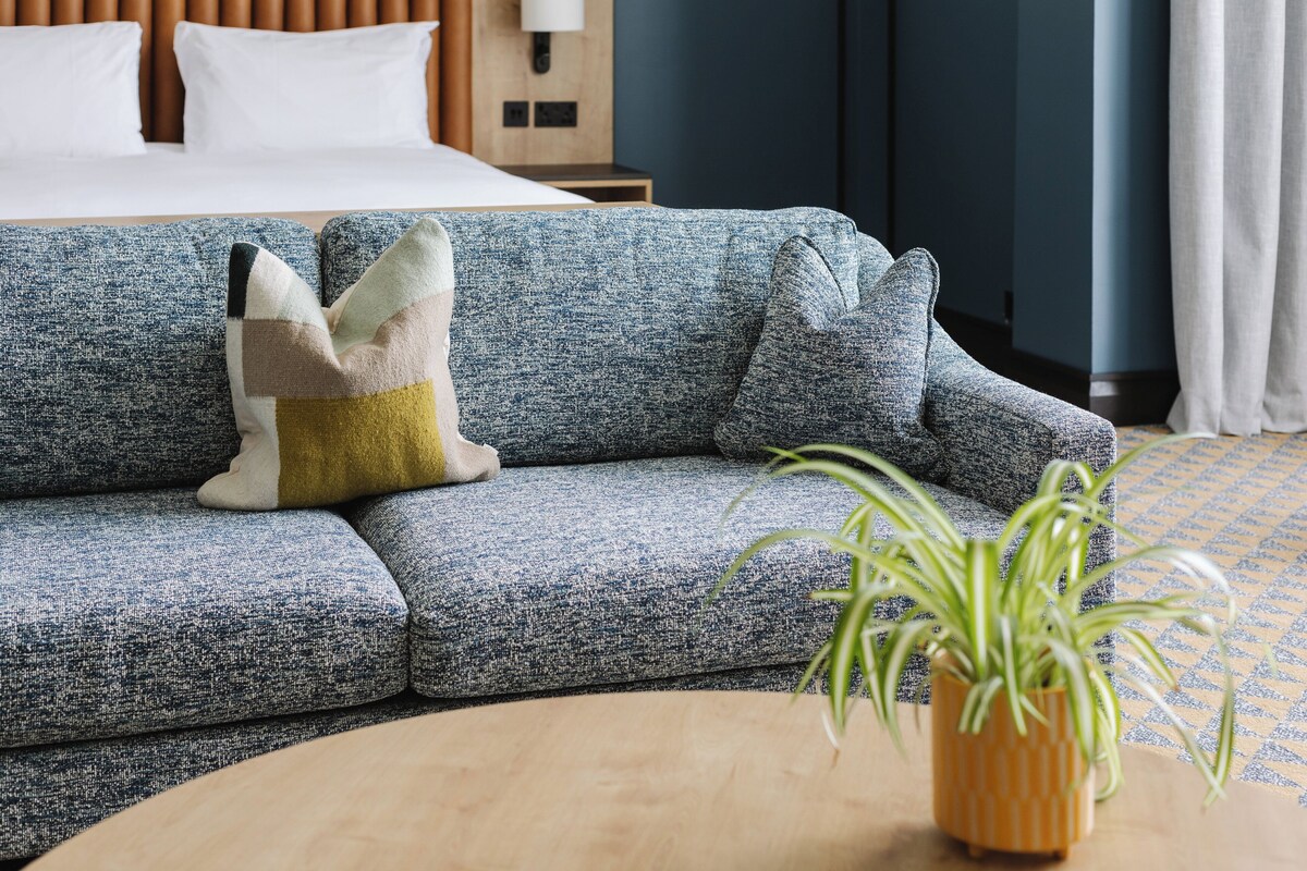 Een moderne hotelkamer met een blauwe gestructureerde bank, een ronde houten salontafel en een potplant.