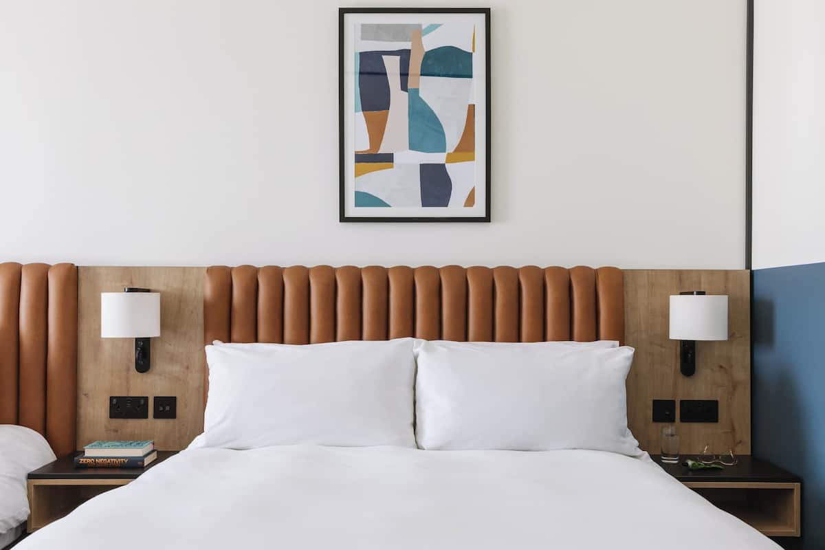 A hotel room with a wooden headboard, white bedding, two bedside tables with lamps, and a framed abstract painting on the wall.