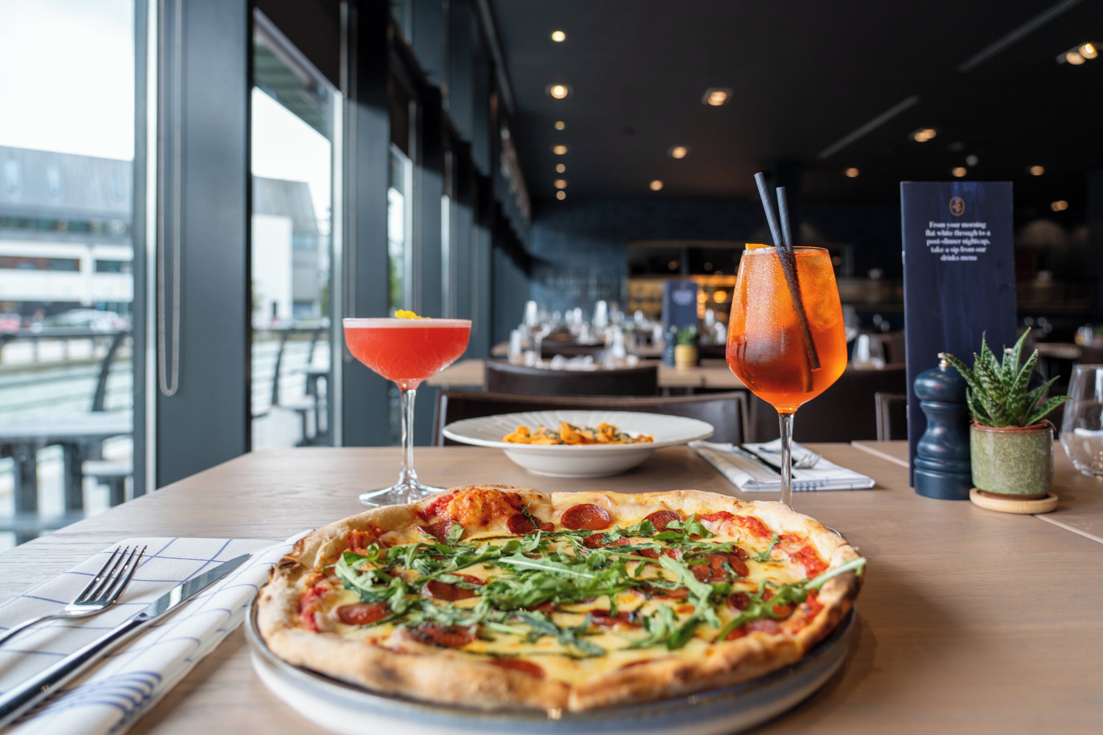 A pizza with toppings and a drink on a table in a restaurant with a view.
