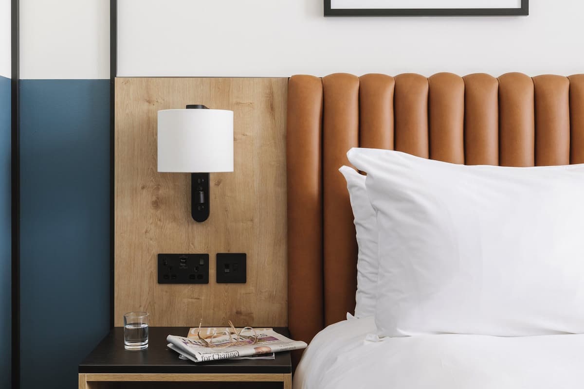 A hotel room with a wooden headboard, a bedside table with a lamp, a magazine, and a glass of water.