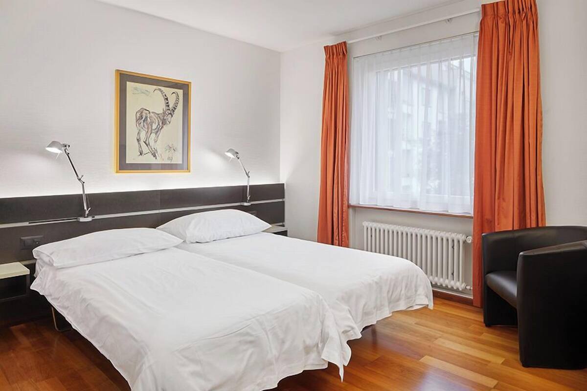 A hotel room with two beds, a black chair, a radiator, and a window with orange curtains.