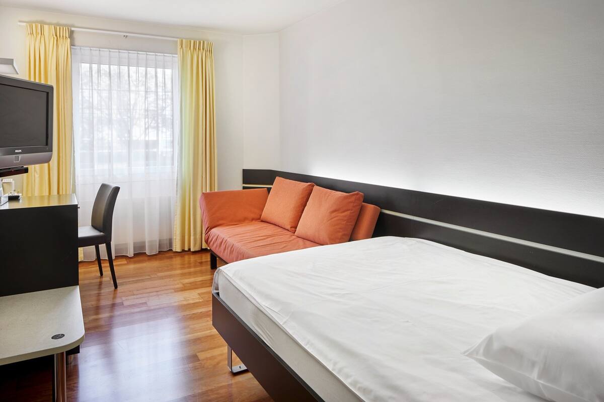 A hotel room with a bed, a desk with a television, and a chair.
