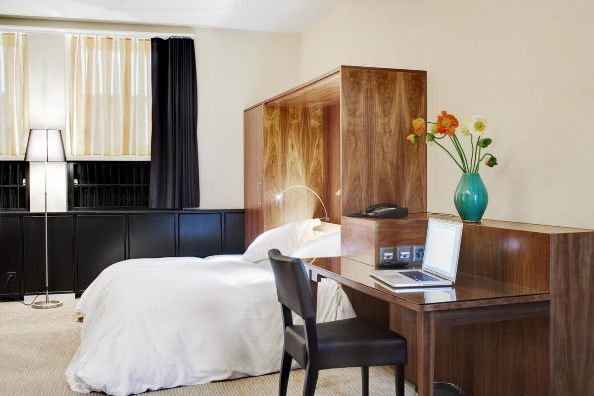 A hotel room with a bed, a desk with a laptop, a telephone, a vase with flowers, and a floor lamp.