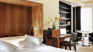 Ein Hotelzimmer mit Holz Kopfteil, einem Schreibtisch mit Laptop, einer Blumenvase, einem Stuhl und einem kleinen Tisch mit Obstschale.