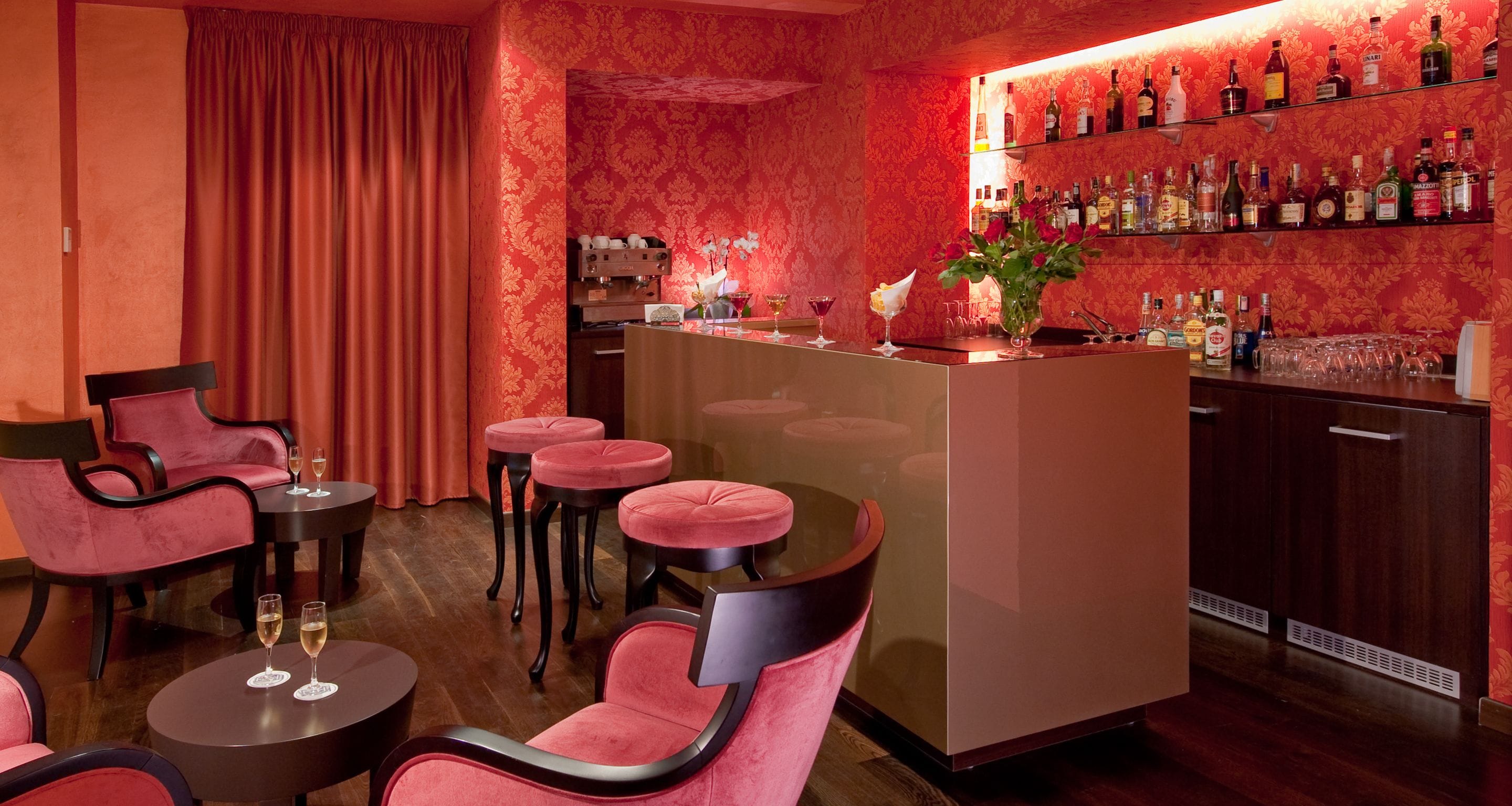 A bar area with a red and gold patterned wall, a bar counter, and several stools.
