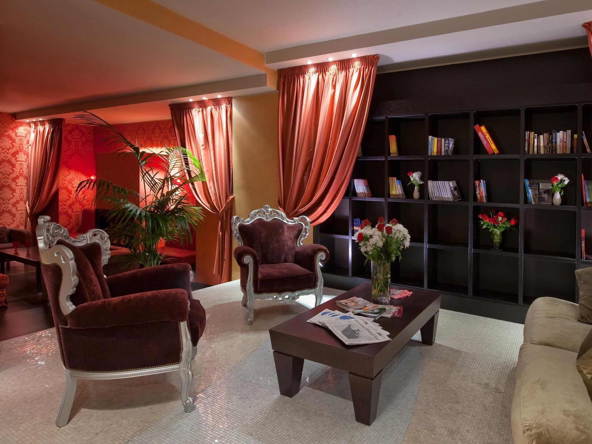 A living room with two ornate armchairs, a coffee table with newspapers, a bookshelf, and a potted plant.