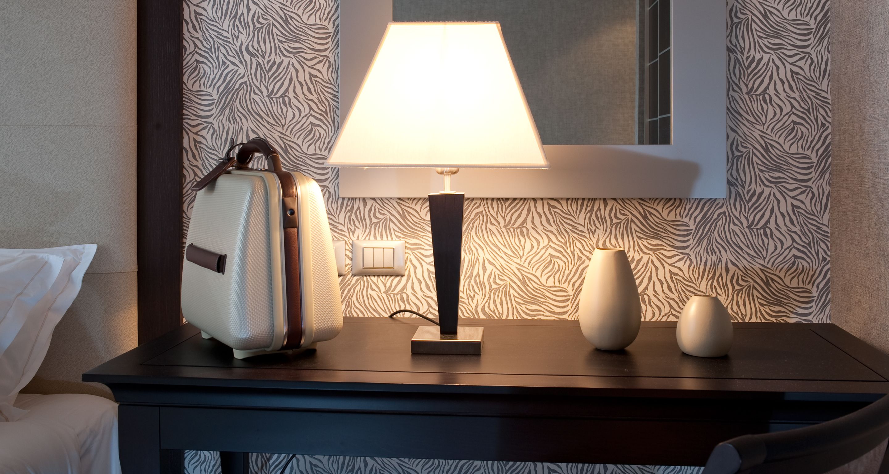 A hotel room with a bed, a bedside table with a lamp, a suitcase, and a vase.