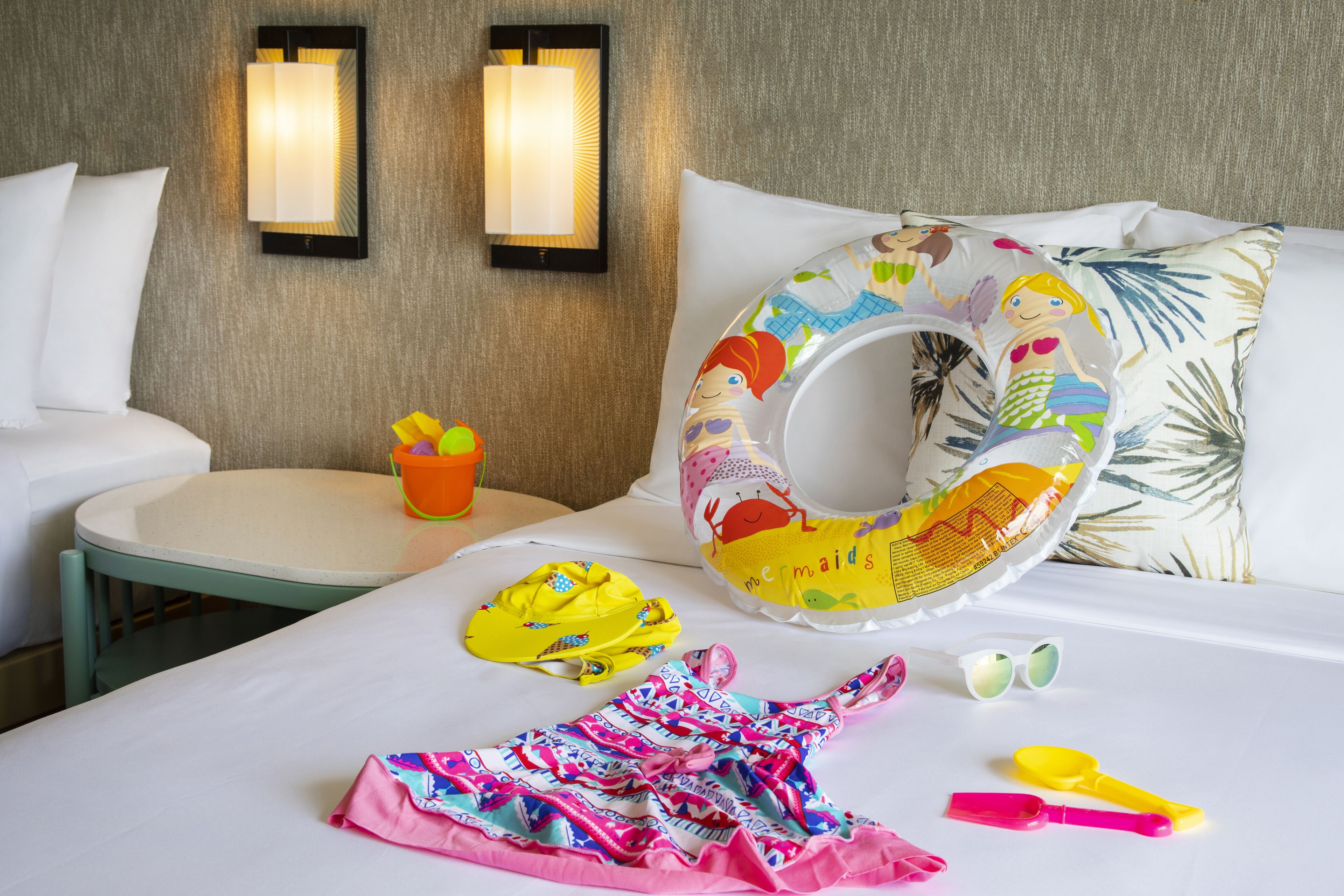 A hotel room with a bed, a small table with a bucket and a bottle, and a colorful mermaid-themed pillow.