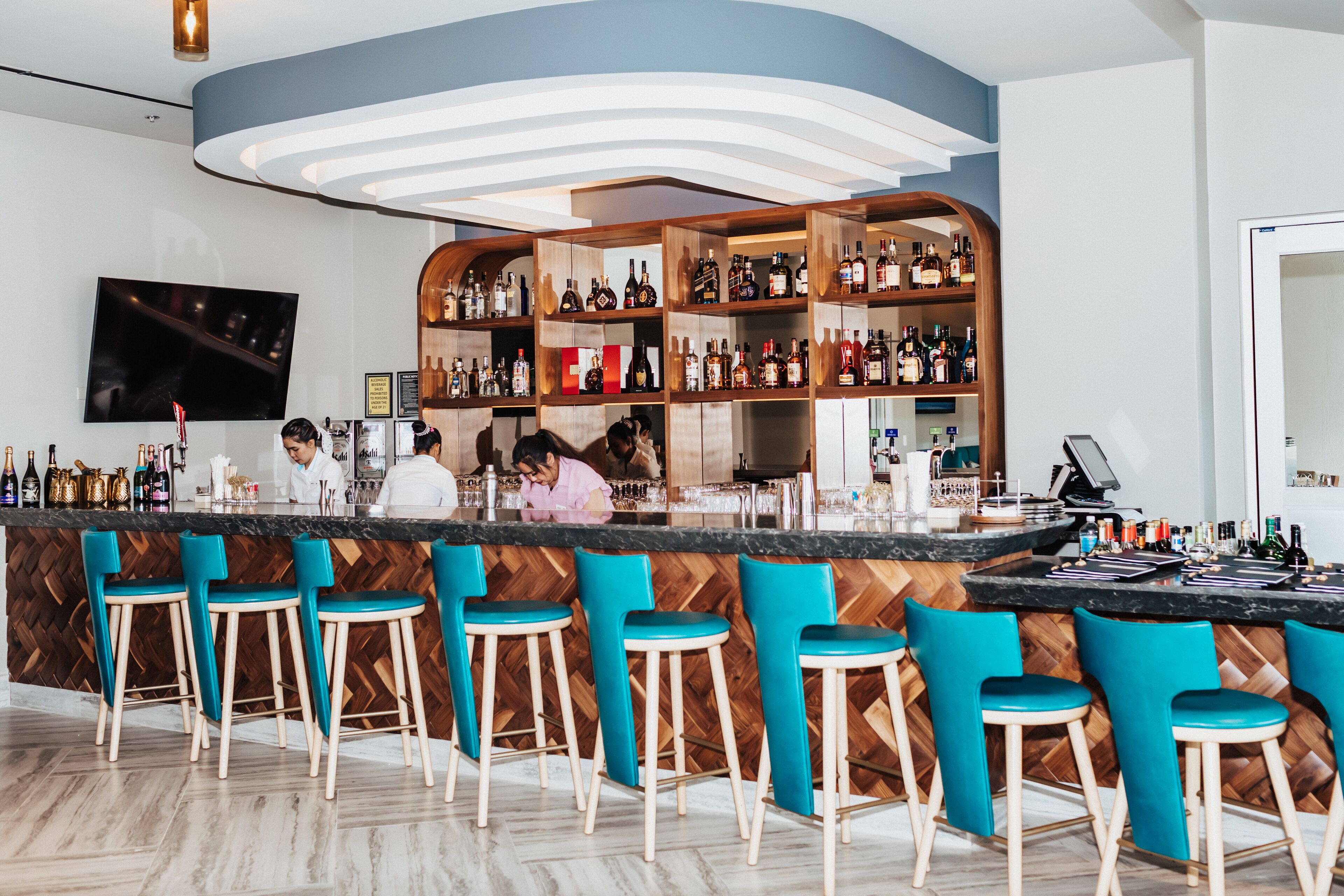 A bar area with a wooden herringbone floor, teal bar stools, and a variety of liquor bottles on shelves.