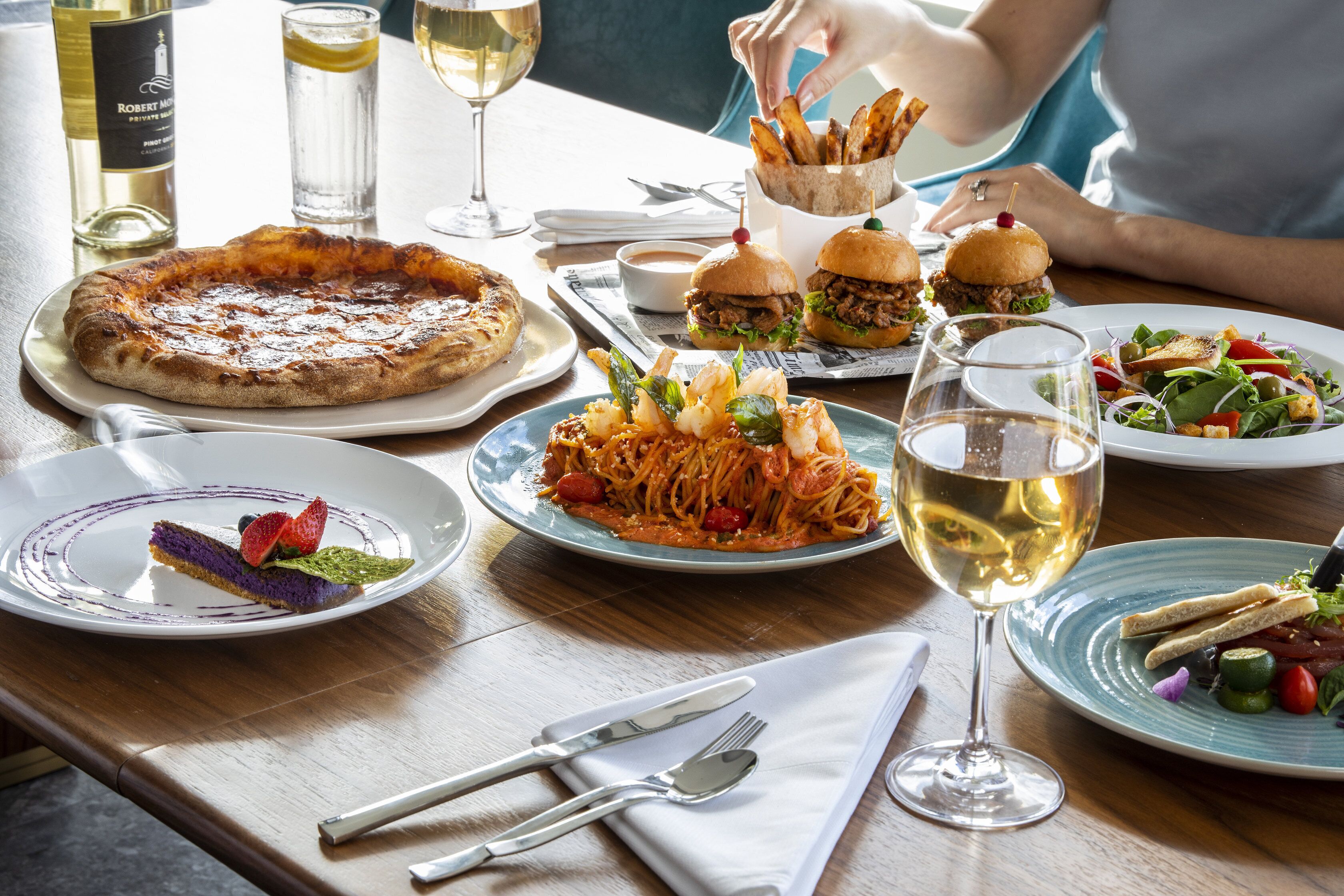 Ein Esstisch mit verschiedenen Gerichten, darunter Pizza, Pasta, Burger und Salat, sowie Weingläsern und Besteck.