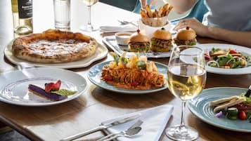 A dining table with various dishes, including pizza, pasta, burgers, and a salad, along with wine glasses and cutlery.