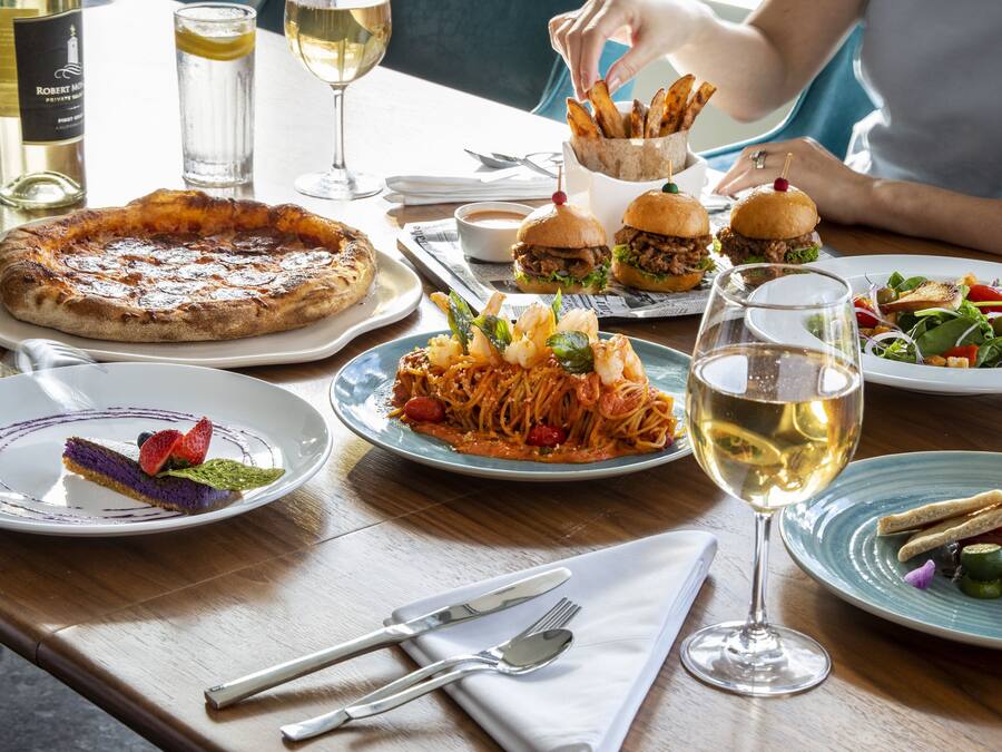 A dining table with various dishes, including pizza, pasta, burgers, and a salad, along with wine glasses and cutlery.