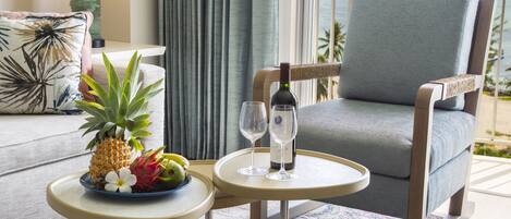 A room with a view of the ocean, a dining table with a plate of fruit and two wine glasses, and a comfortable chair.