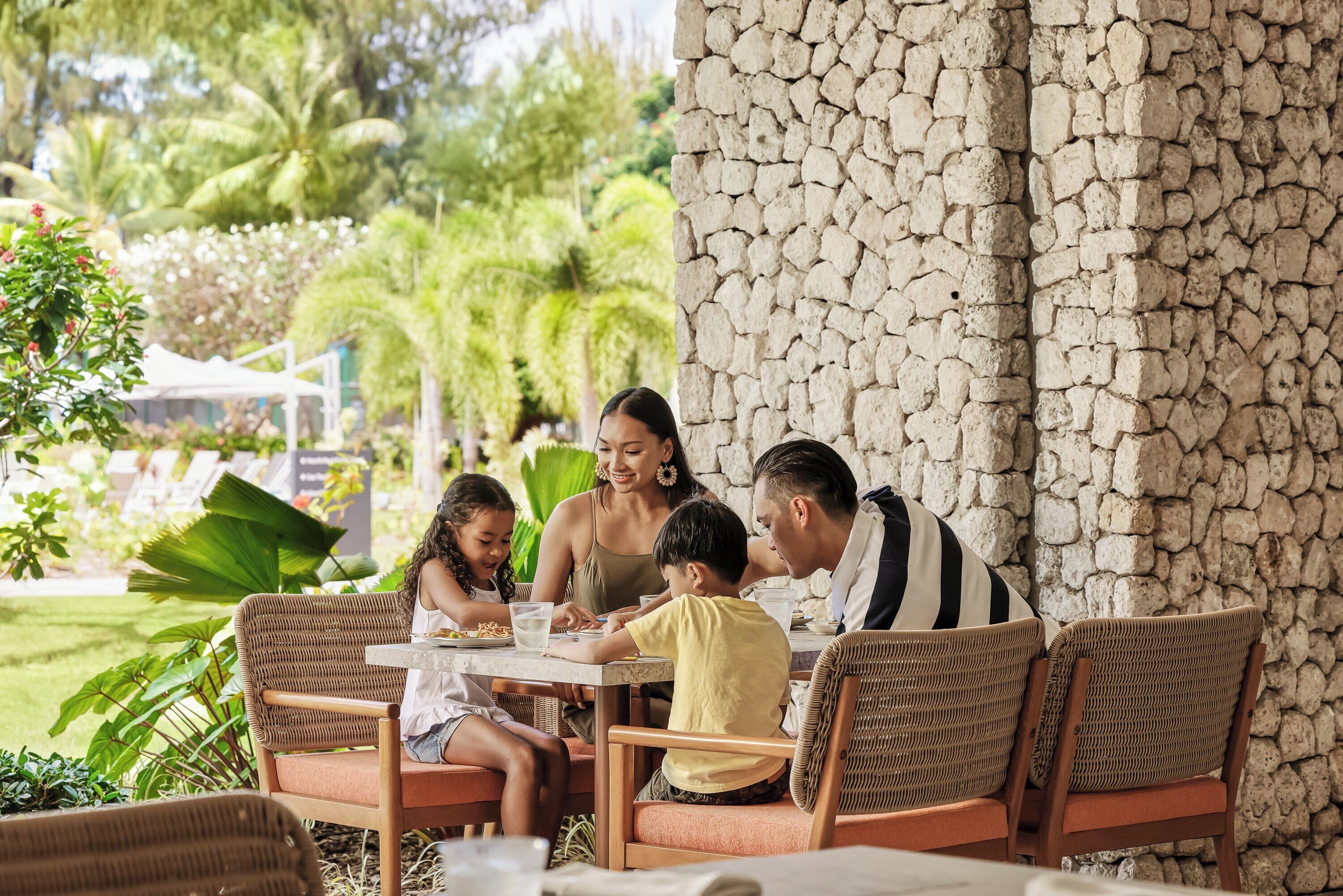 Une famille est assise à une table à l’extérieur, autour de chaises en osier, avec un mur de pierre en arrière-plan.