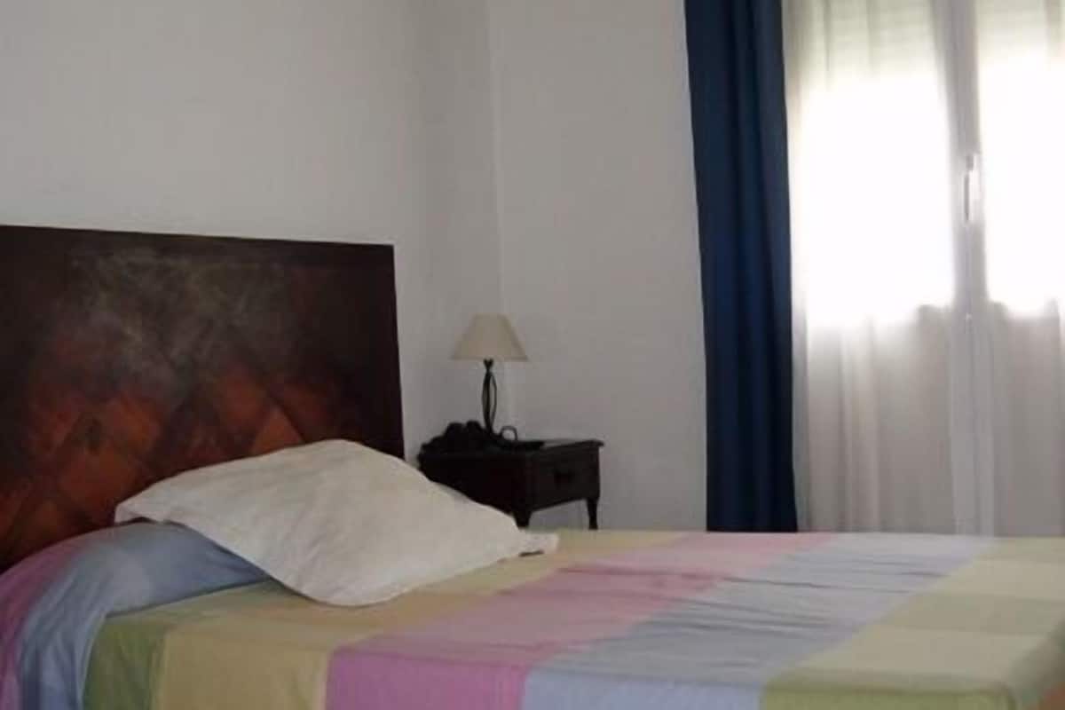 Un dormitorio con cama, mesita de noche, lámpara y ventana con cortinas.