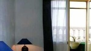 Un dormitorio con cama, mesita de noche, lámpara, ventana con cortinas y vistas al exterior.