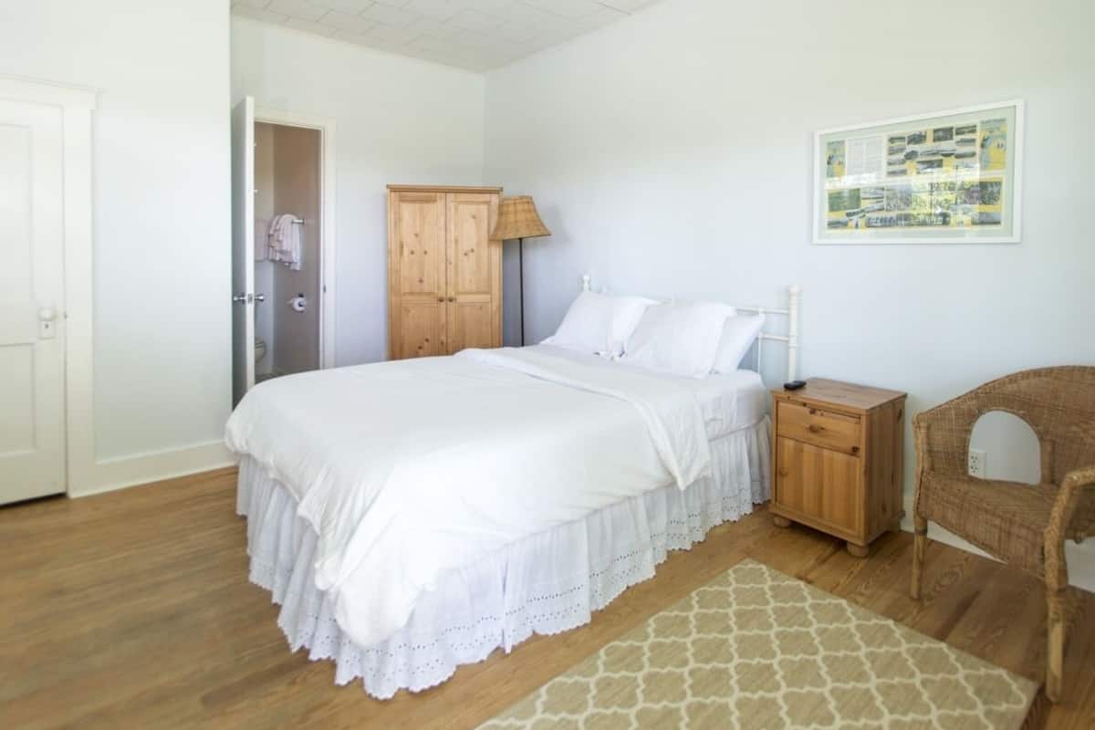 A bedroom with a bed, a wooden nightstand, a wicker chair, a wooden wardrobe, and a framed picture on the wall.