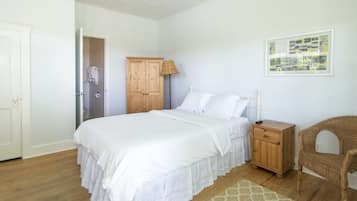 A bedroom with a bed, a wooden nightstand, a wicker chair, a wooden wardrobe, and a framed picture on the wall.