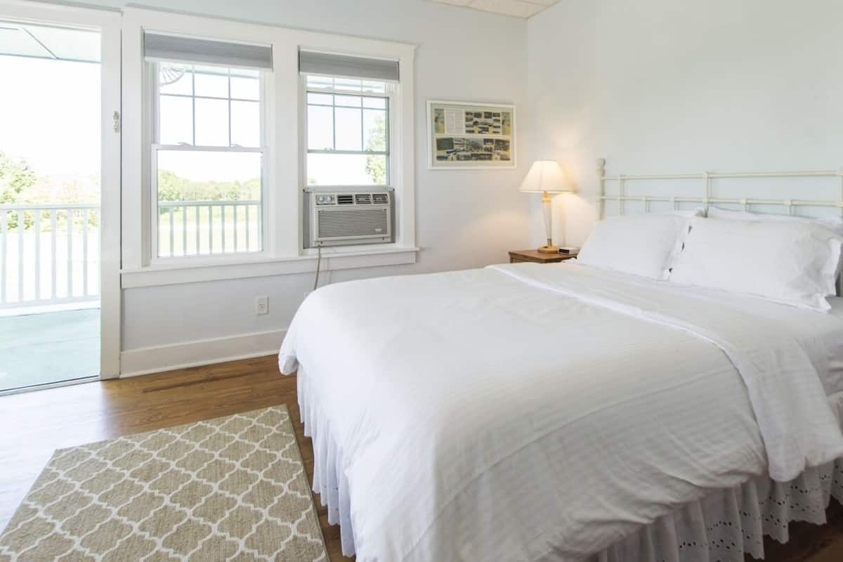 A bedroom with a bed, a nightstand, a lamp, a window with blinds, and a wall-mounted air conditioner.