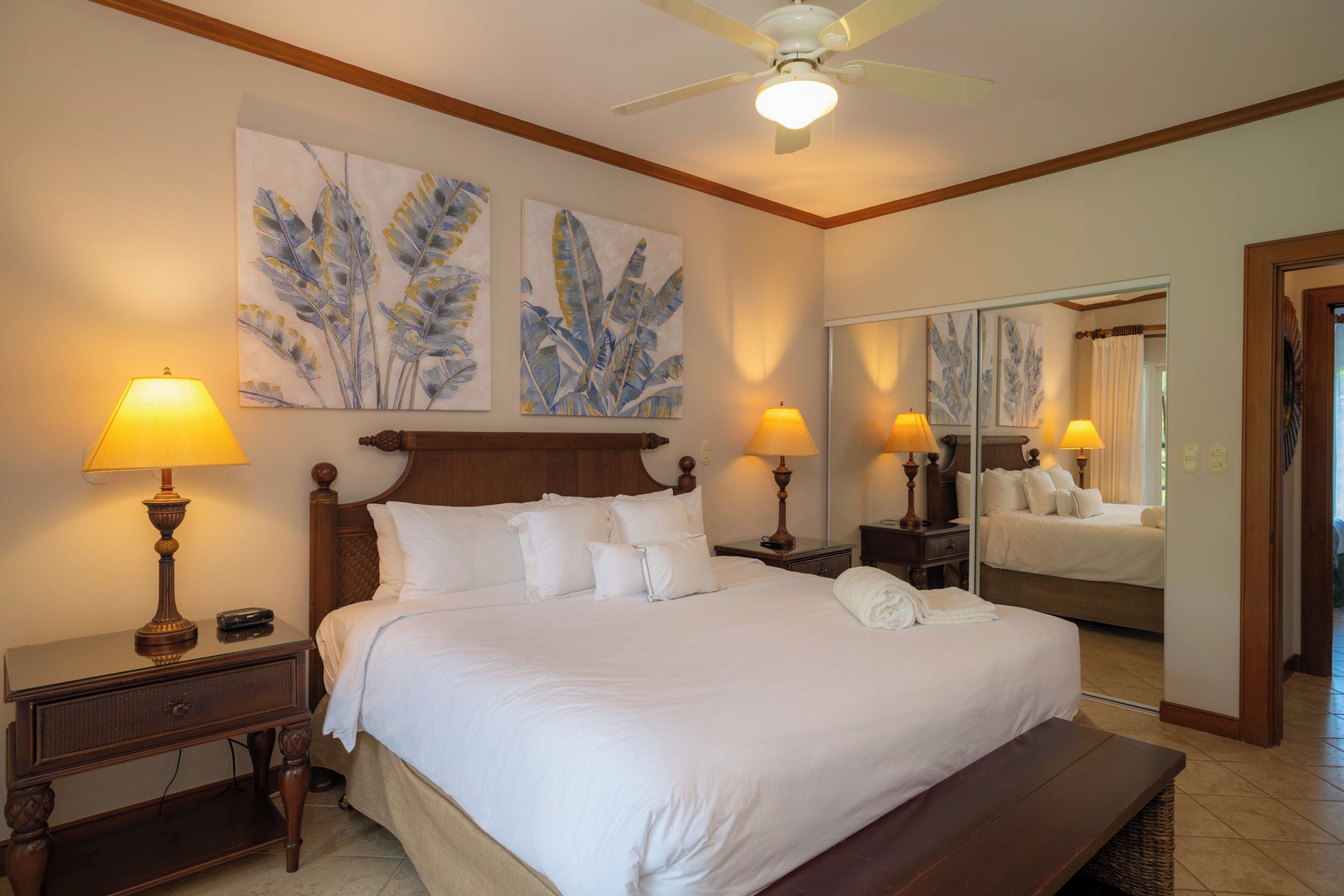 A hotel room with a large bed, two bedside lamps, a ceiling fan, and artwork on the walls.