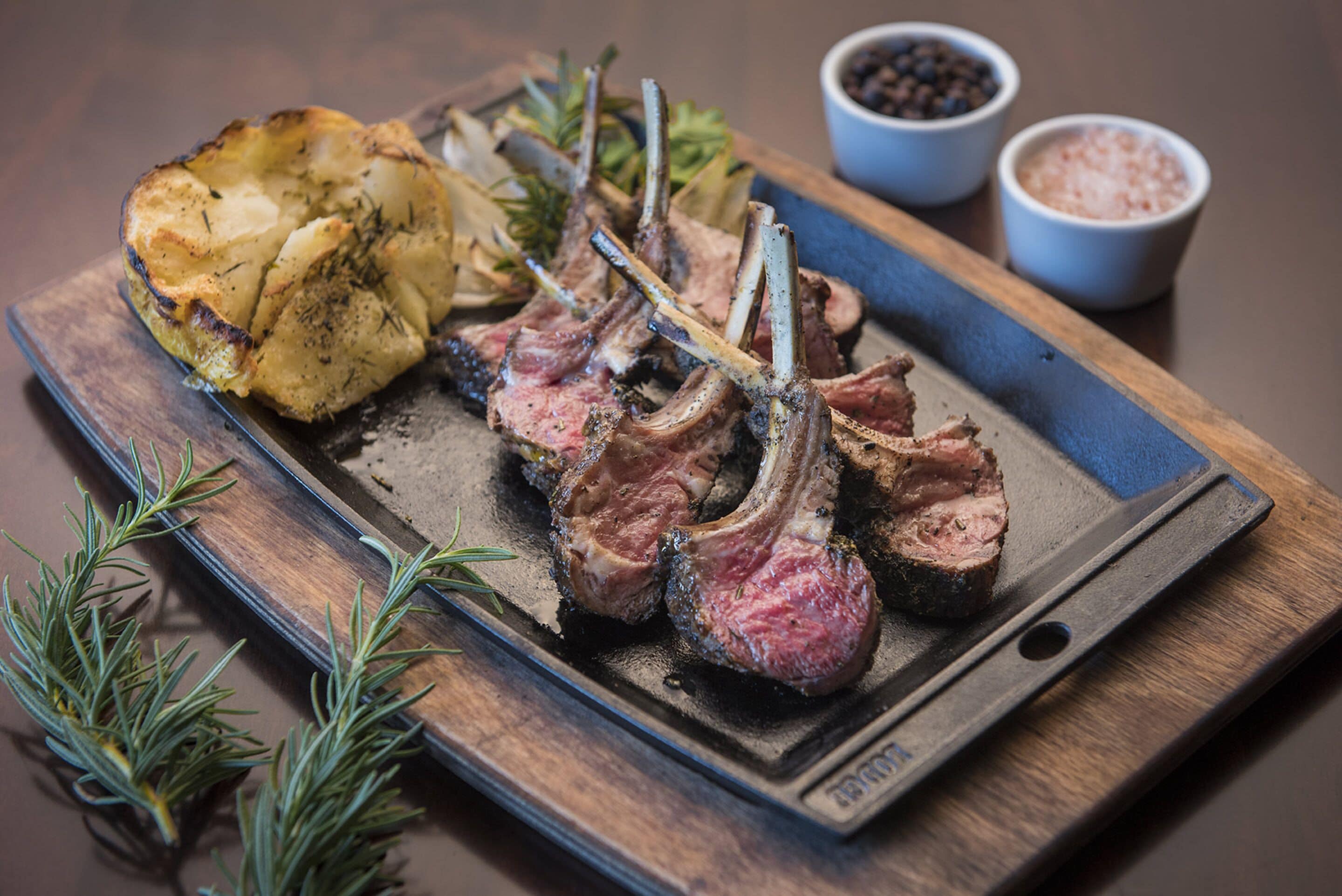 A wooden serving board with grilled lamb chops, roasted potatoes, and rosemary sprigs.