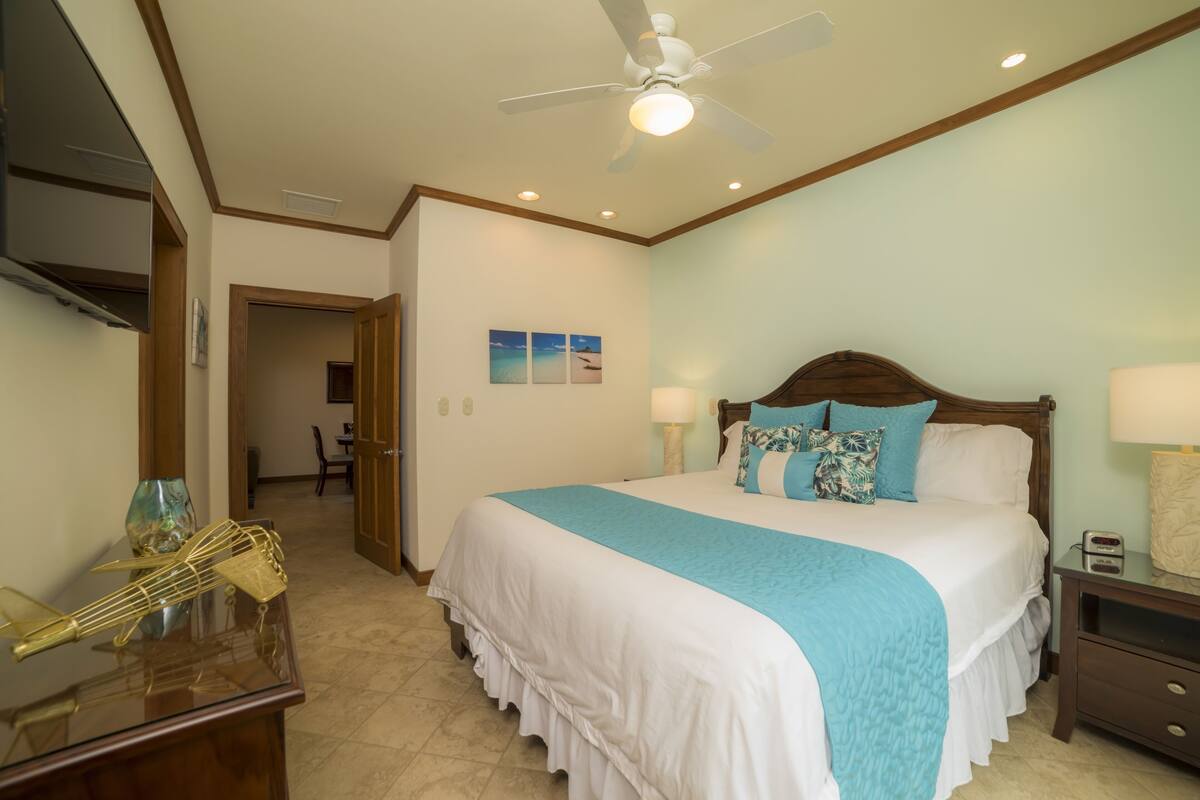 A bedroom with a bed, bedside tables, a ceiling fan, and a view of the hallway.