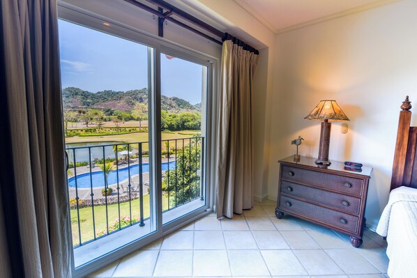 Una habitación con un amplio ventanal corredera que da a un área de piscina, un aparador de madera con una lámpara y una cama.