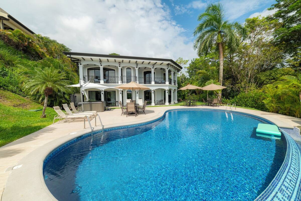 A large, curved swimming pool with lounge chairs and umbrellas, surrounded by lush greenery and a large house with multiple balconies.