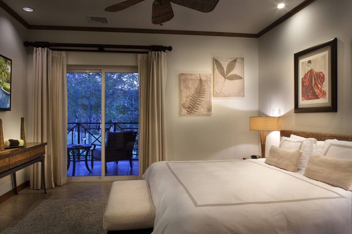 A hotel room with a large bed, a desk, a chair, and a balcony with a view of trees.