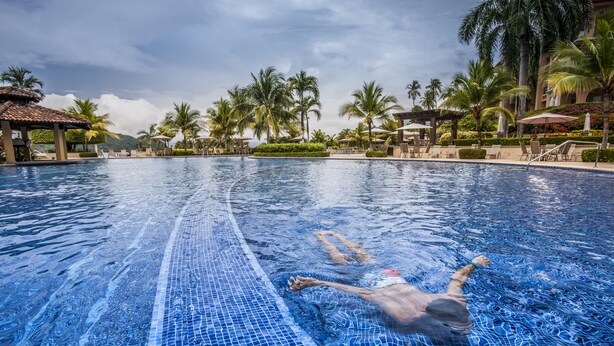 A swimming pool with a tiled edge, surrounded by palm trees and lounge chairs. - Stay In CR Los Sueños Condos (Jacó)