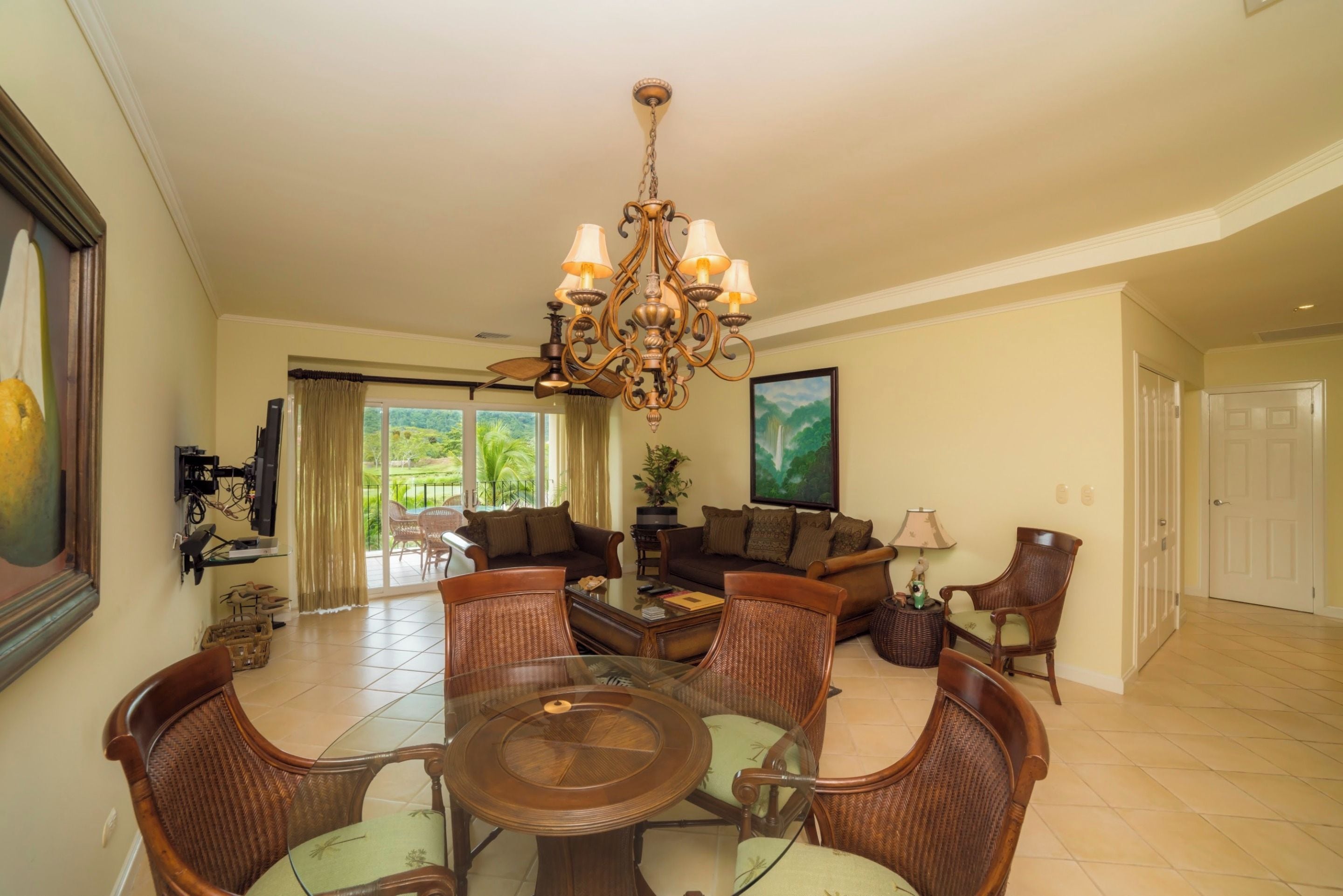 A spacious living room with a chandelier, a dining area with a round table, and a large window with a view of greenery.