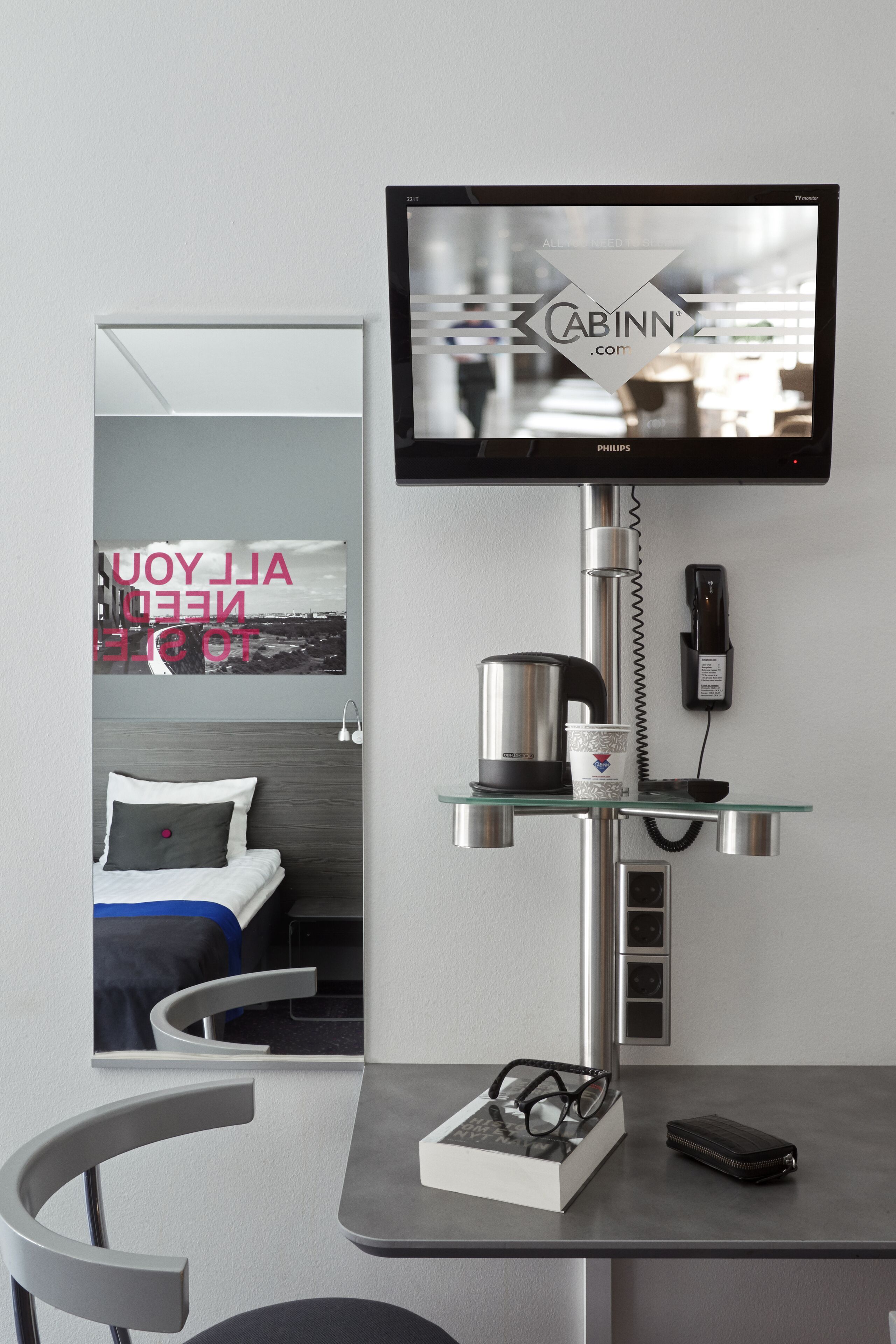 Une chambre d’hôtel moderne dotée d’un téléviseur à écran plat fixé au mur, d’une table de chevet avec une machine à café, d’un téléphone et d’une petite étagère.