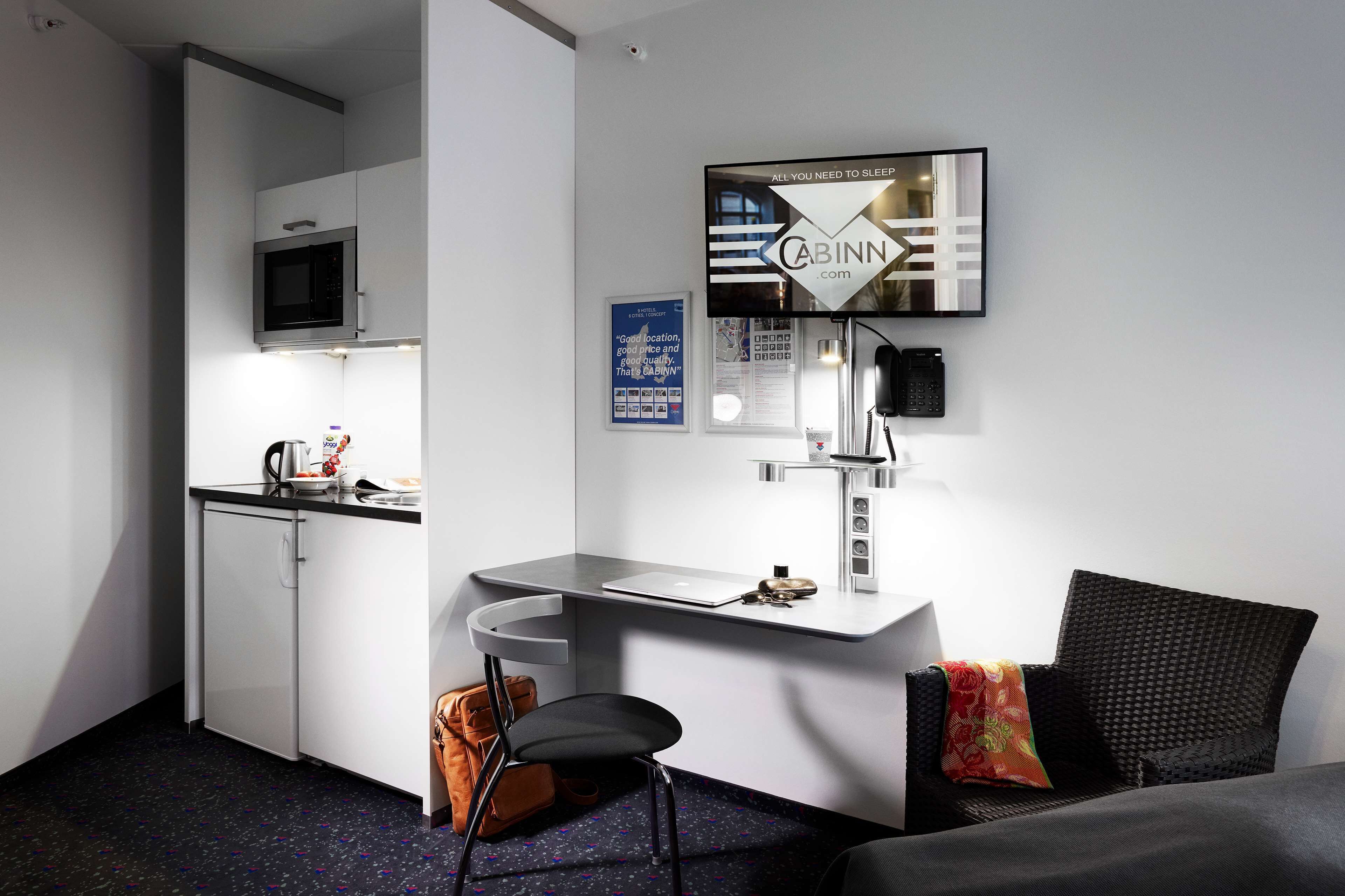 A compact hotel room with a kitchenette, a desk, a chair, a bed, and a TV mounted on the wall.