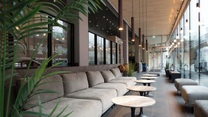 Un espacio interior moderno con sofás, mesas redondas, ventanas amplias y plantas de interior.