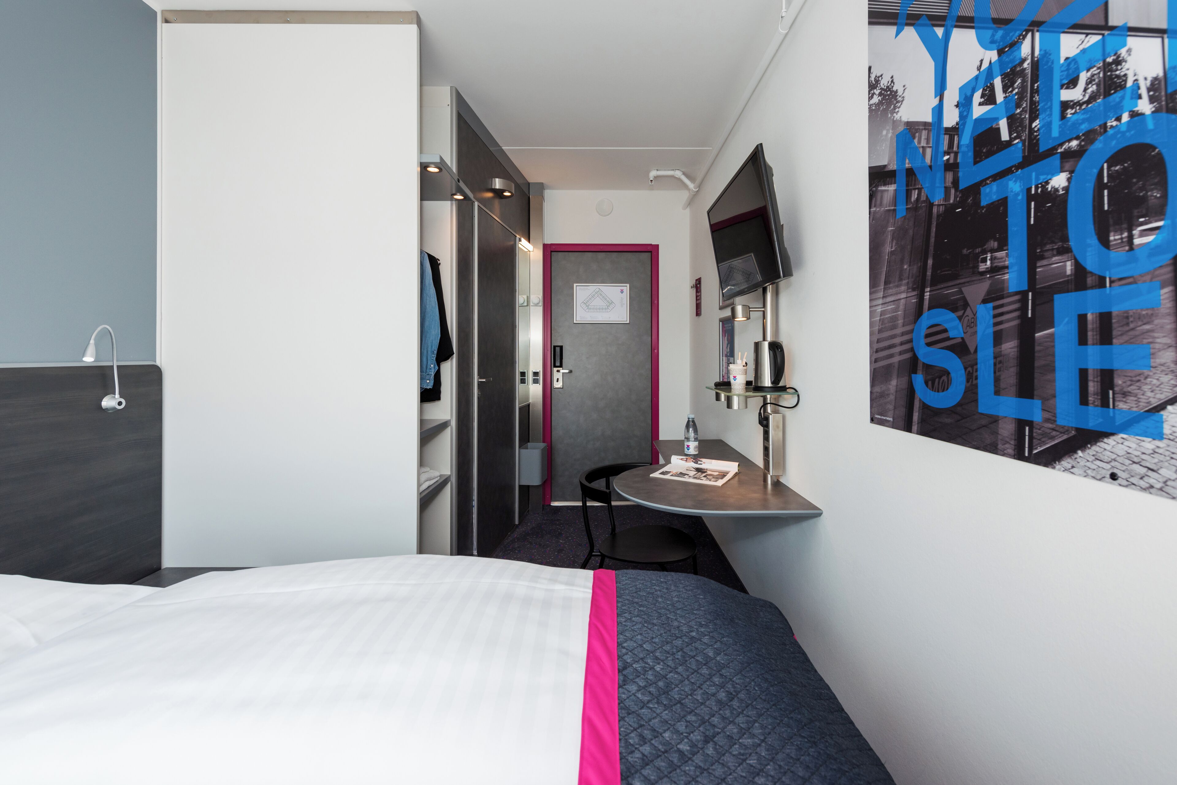 Quarto de hotel com cama, escrivaninha, cadeira, espelho e porta.