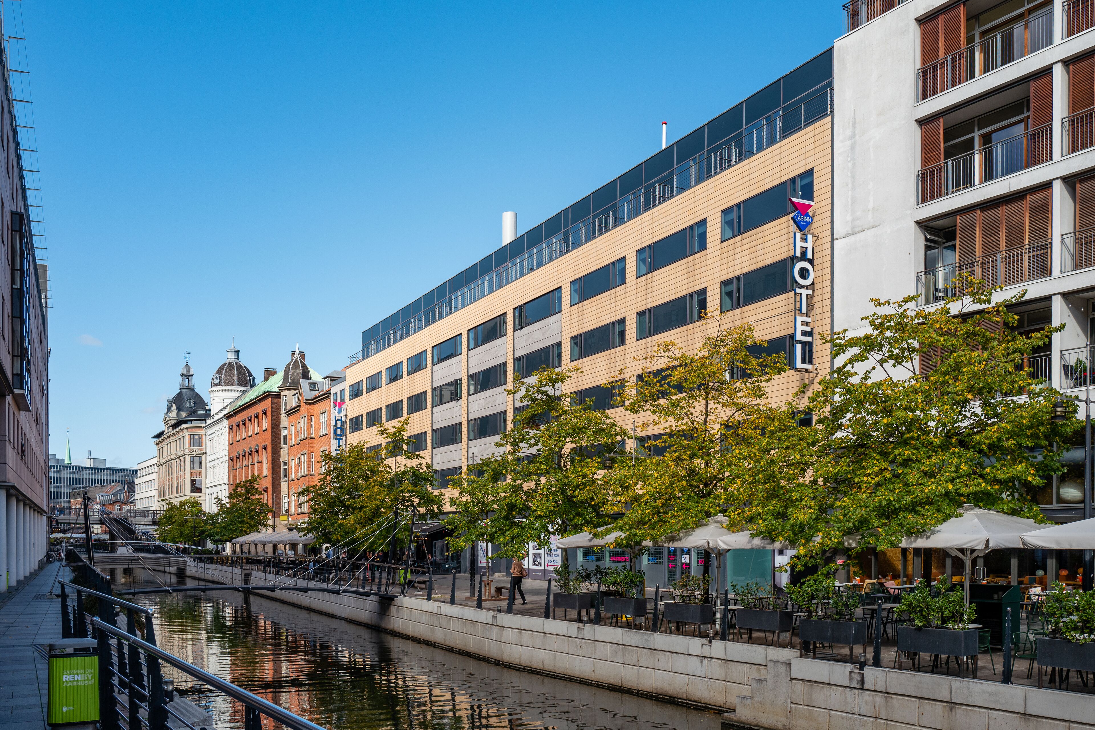 A hotel with a modern facade, a river in the foreground, and outdoor seating areas.