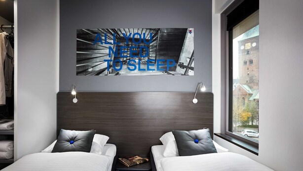 Una habitación de hotel moderna con dos camas, un cabecero con una obra de arte de un paisaje urbano, una mesita de noche con una lámpara y una ventana que da a vistas de edificios y un coche.