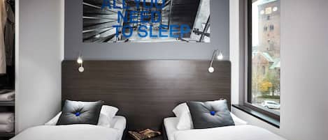 A modern hotel room with two beds, a headboard with a cityscape artwork, a small table with a lamp, and a window with a view of buildings and a car.