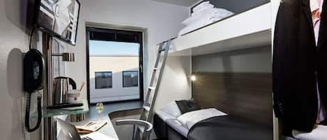 A compact hotel room with a bunk bed, a desk with a laptop, a chair, a small table with a wine glass, a telephone, and a window with a view of buildings.
