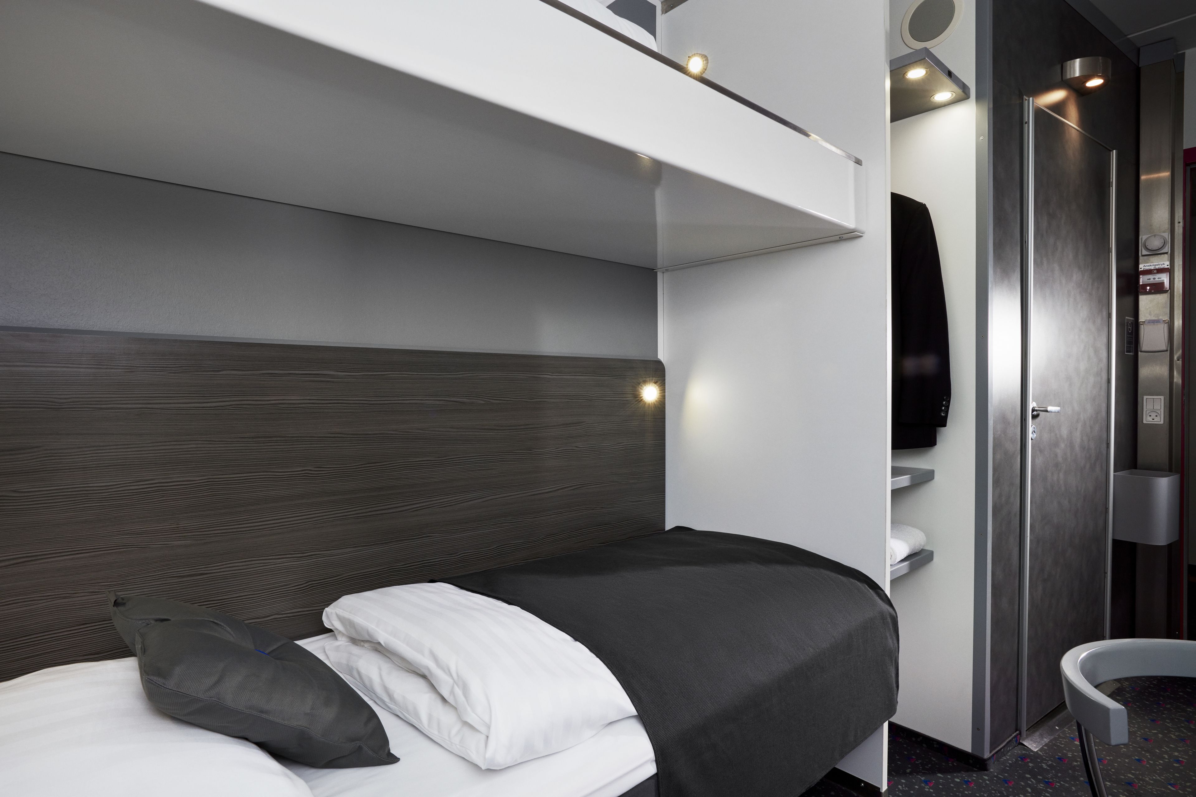Una habitación de hotel moderna con una cama, un banco empotrado, un pequeño rincón de descanso y un baño con espejo y lavamanos.