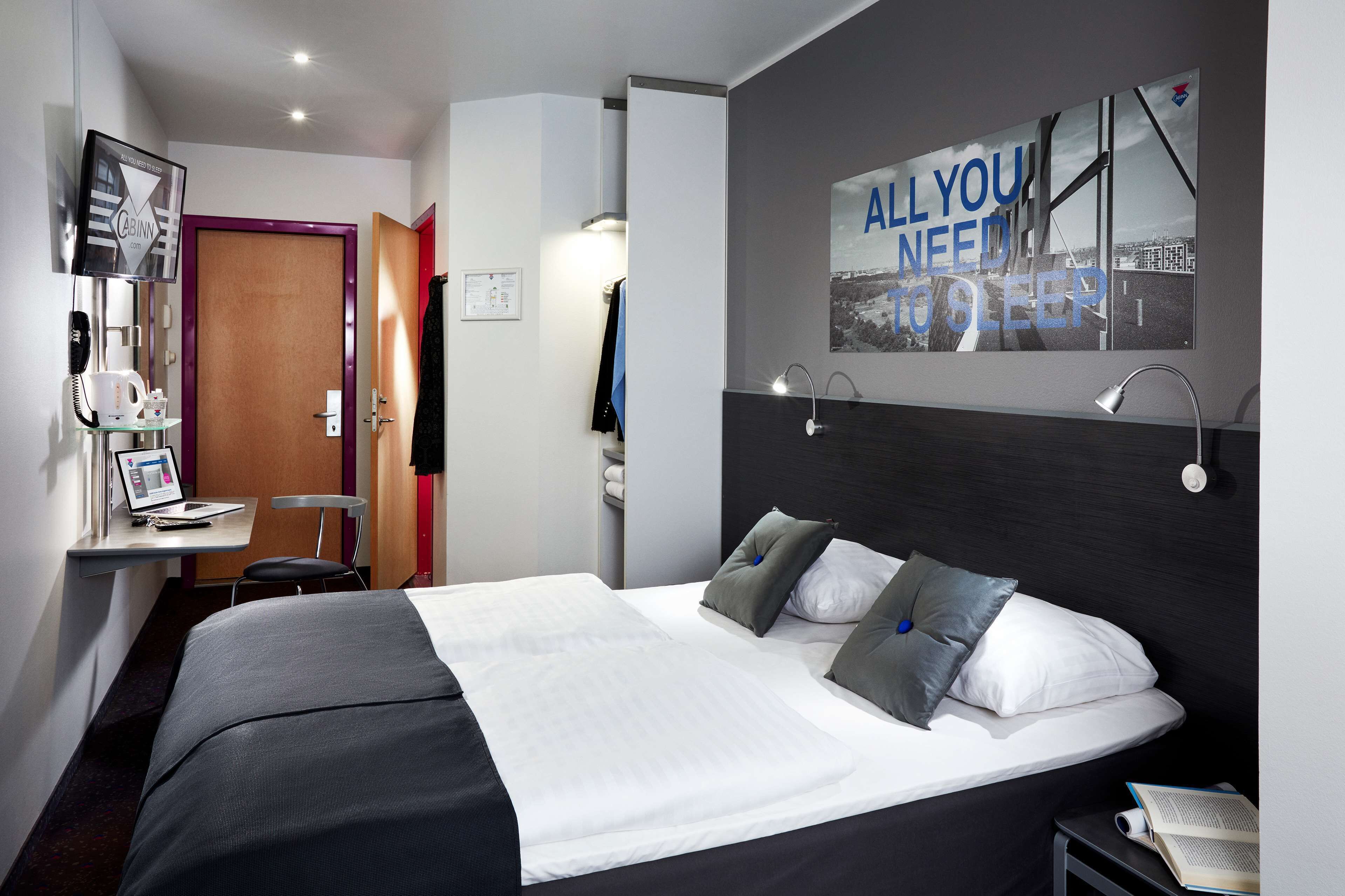 A modern hotel room with a large bed, a desk with a computer, a chair, and a view of the city through the window.