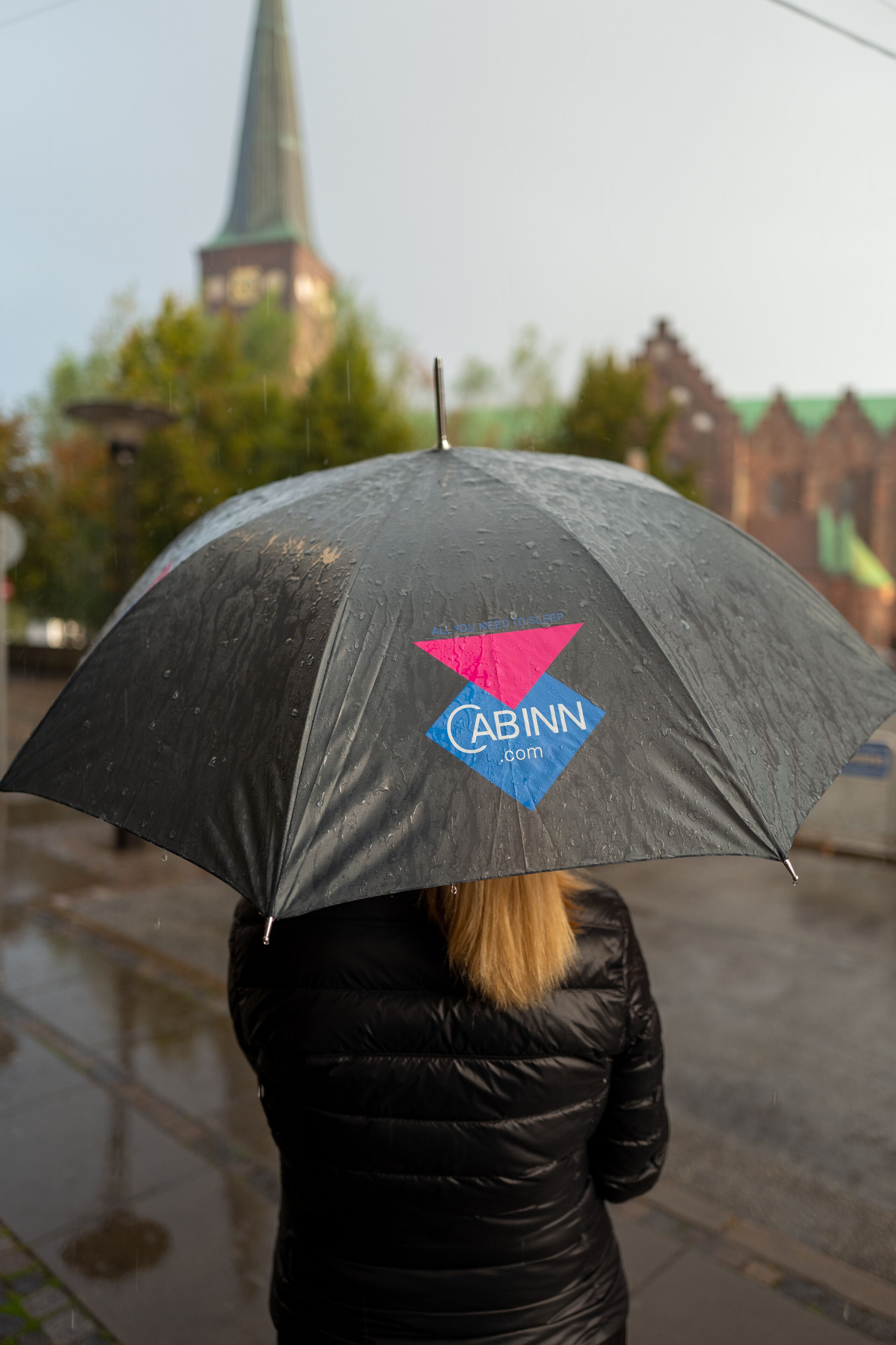 A person holding a black umbrella with a logo, standing in a rainy outdoor setting.