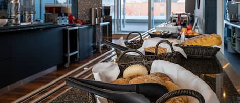A breakfast buffet with various breads and pastries, a coffee station, and a view of the outside through large windows.