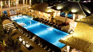 A large, rectangular swimming pool with a thatched-roof structure adjacent to it, surrounded by palm trees and well-lit by ambient lighting. - Riyuegu Hotsprings Resort (Xiamen)