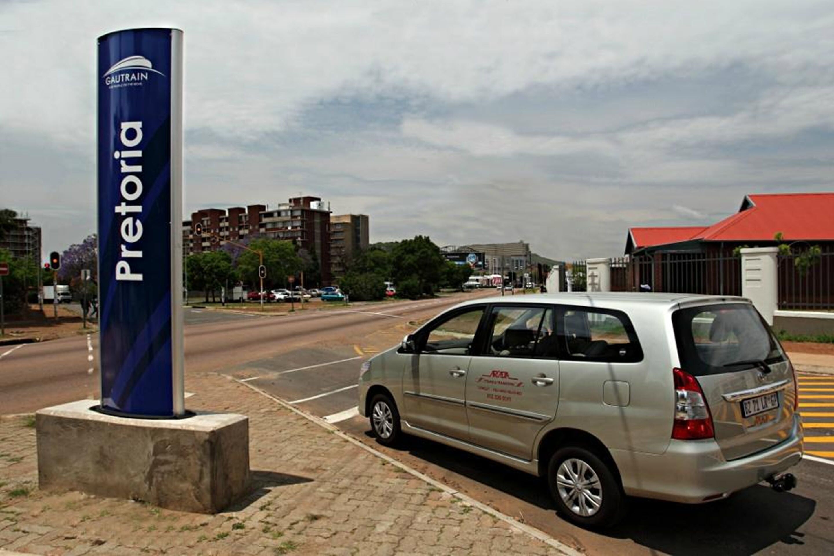 Una minivan plateada estacionada cerca de un cartel azul con la palabra 'Pretoria' escrita.