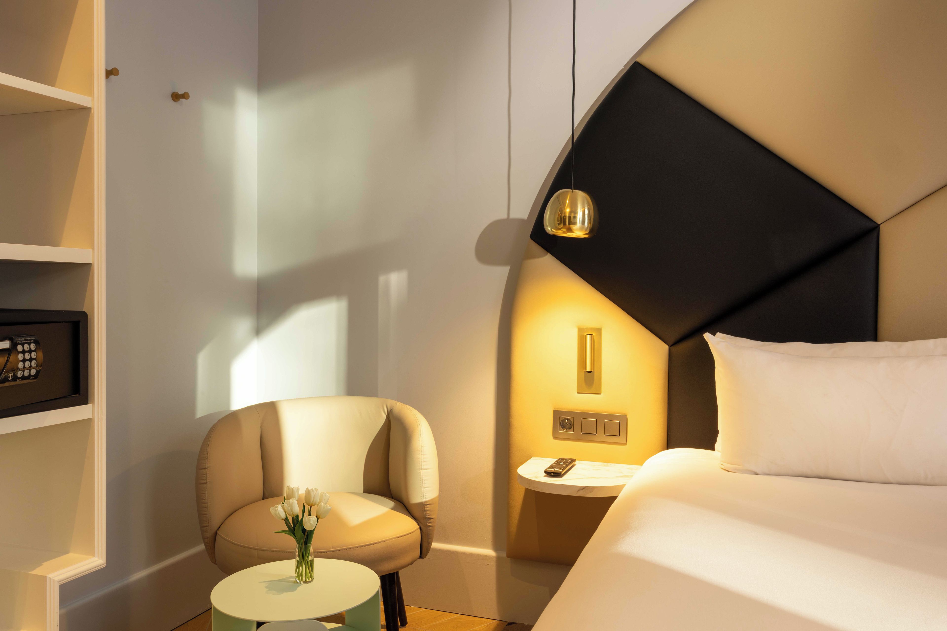 A hotel room with a bed, a chair, a small table with a vase of flowers, a bedside table with a lamp, and a wall-mounted control panel.